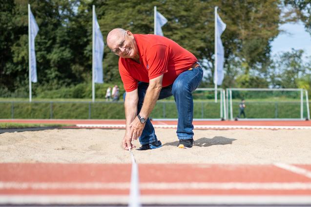 Mann in rotem Shirt richtet eine Messleine auf einem Sandplatz in einem Sportstadion aus.